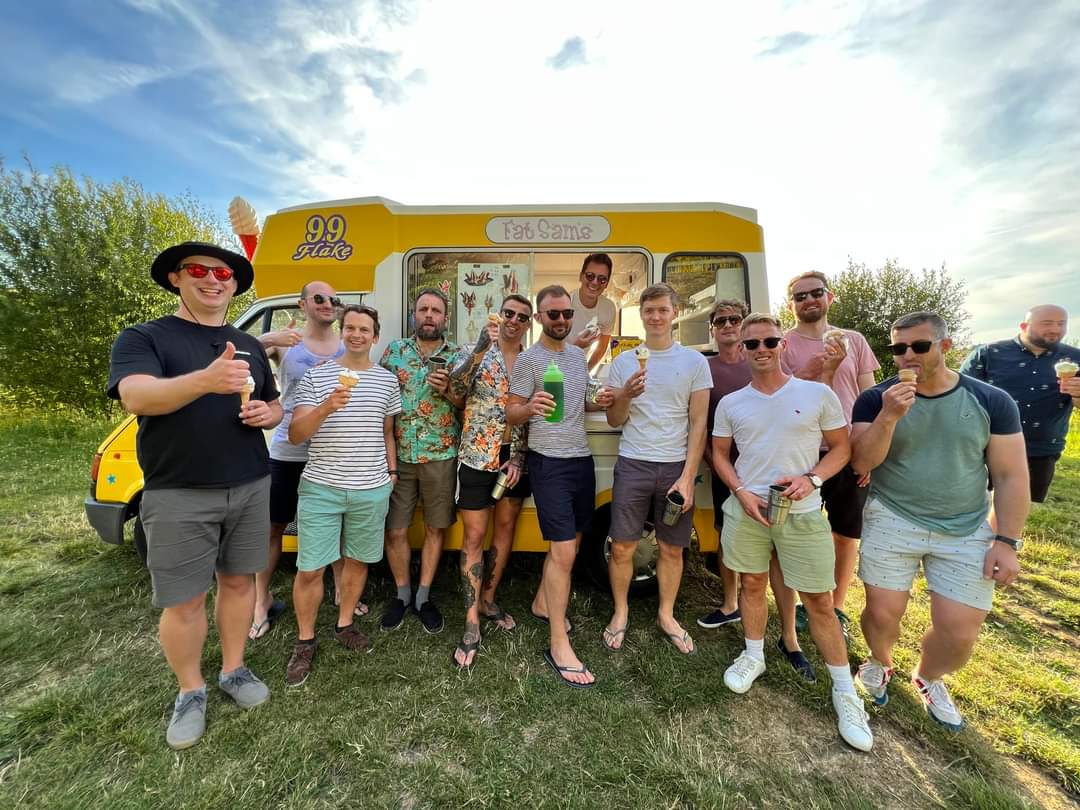 A group of cheerful men posing in front of a yellow "Fat Sam's" ice cream van on a sunny day. They are smiling, enjoying ice cream cones, and holding drinks, creating a fun and relaxed atmosphere. The van's "99 Flake" branding and menu board are visible, and the backdrop features green trees and a bright sky, adding to the vibrant outdoor setting.