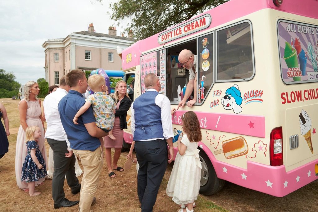 A lively wedding scene featuring a group of guests gathered around a pink and cream vintage ice cream van decorated with playful illustrations and text, including "Try our slush" and "Enjoy the best!" Guests of all ages, including children, are excitedly waiting to be served, with a server leaning out of the van's window to hand out treats. The background includes a grand wedding venue and an inflatable play structure, adding to the festive and joyful atmosphere.