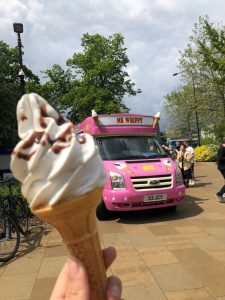 A close-up of a hand holding a soft-serve vanilla ice cream cone drizzled with chocolate sauce, with a pink "Mr Whippy" ice cream van visible in the background. The van is parked on a paved area surrounded by lush green trees under a cloudy sky. The vibrant colours and cheerful setting create a fun and inviting atmosphere.