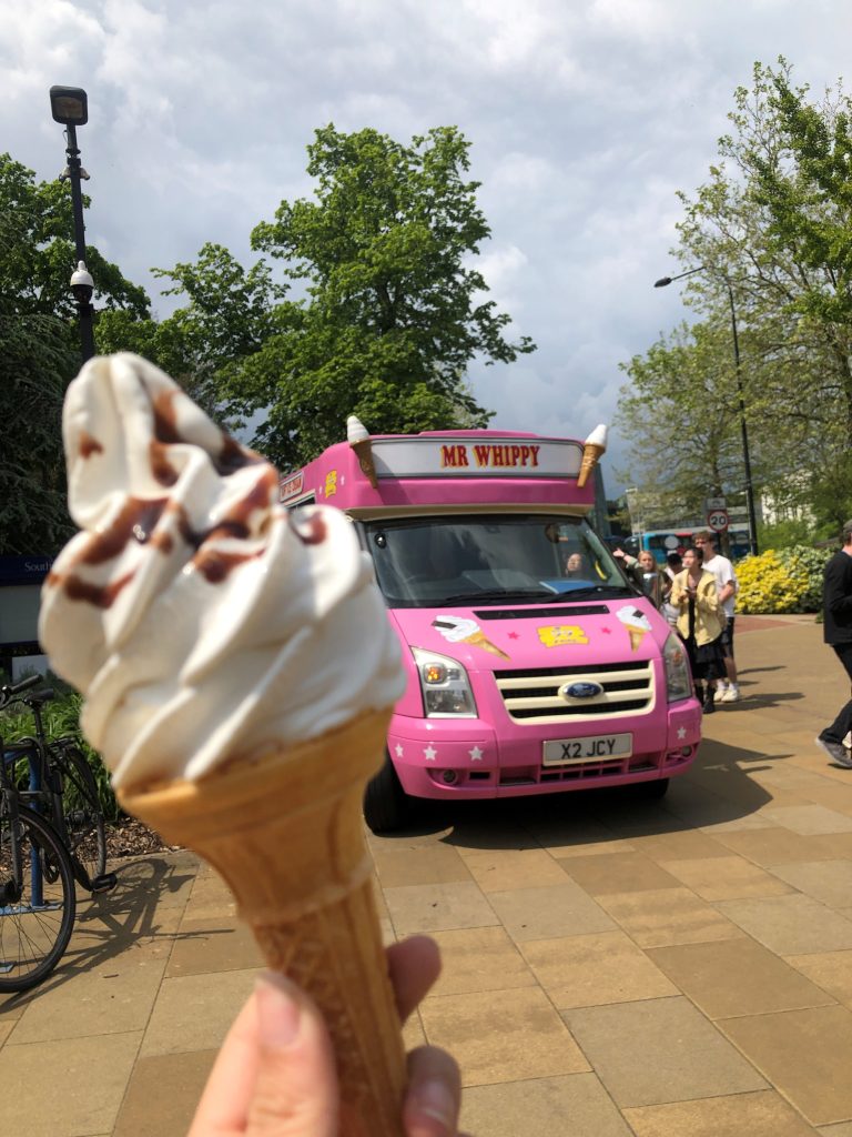 A close-up of a hand holding a soft-serve vanilla ice cream cone drizzled with chocolate sauce, with a pink "Mr Whippy" ice cream van visible in the background. The van is parked on a paved area surrounded by lush green trees under a cloudy sky. The vibrant colours and cheerful setting create a fun and inviting atmosphere.