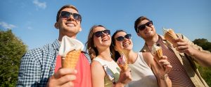 A group of four friends standing outdoors on a sunny day, smiling and enjoying ice cream cones. They are all wearing sunglasses and casual summer outfits, with a bright blue sky and greenery in the background, creating a cheerful and vibrant atmosphere.