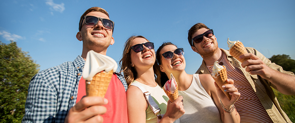 A group of four friends standing outdoors on a sunny day, smiling and enjoying ice cream cones. They are all wearing sunglasses and casual summer outfits, with a bright blue sky and greenery in the background, creating a cheerful and vibrant atmosphere.