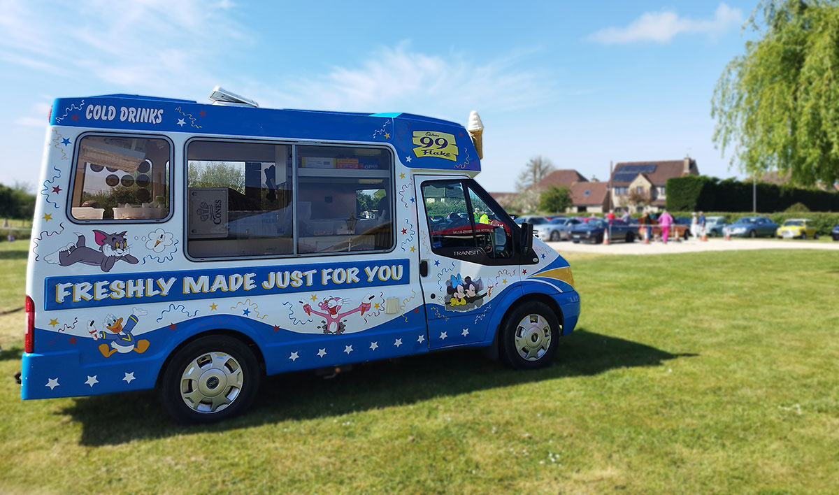 One of Fat Sam's Ice Creams bright blue ice cream vans parked on a neatly mowed grassy field at a vintage vehicle meeting in Wiltshire. The van is decorated with colourful cartoon characters, including Tom from Tom and Jerry, Donald Duck, and Mickey Mouse. Bold text on the side reads "Freshly Made Just For You" and "99 Flake," with stars and playful designs adding to the cheerful look. The background features a sunny blue sky, a few wispy clouds, and a line of cars and houses, with people milling about, creating a lively, family-friendly atmosphere.