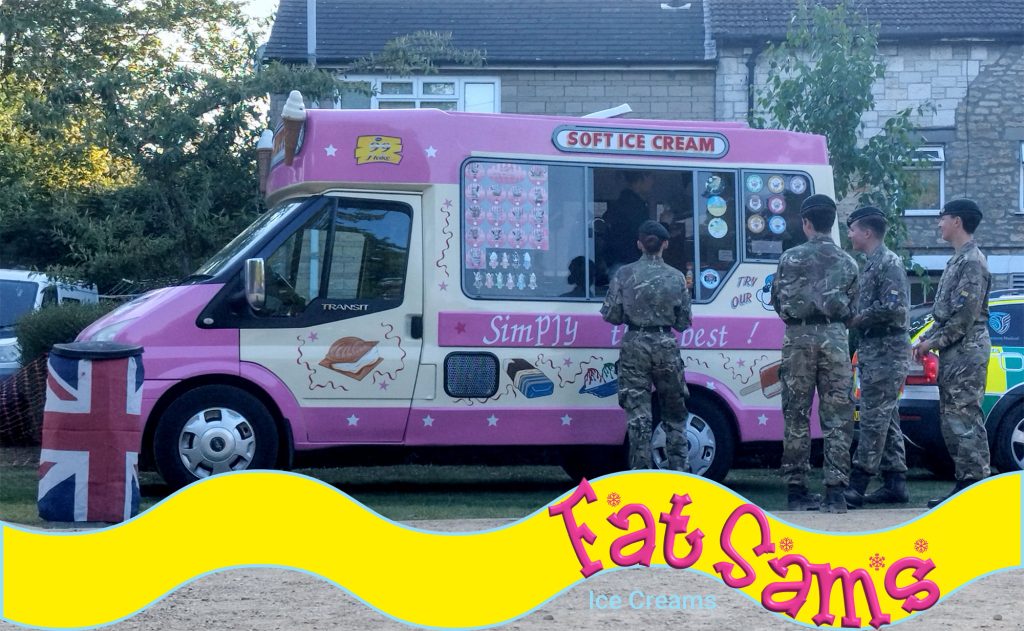 A group of soldiers in camouflage uniforms stand in line at the pink Fat Sam’s van at dusk. An ambulance vehicle is parked nearby, and Union Jack bunting decorates a bin in the foreground.