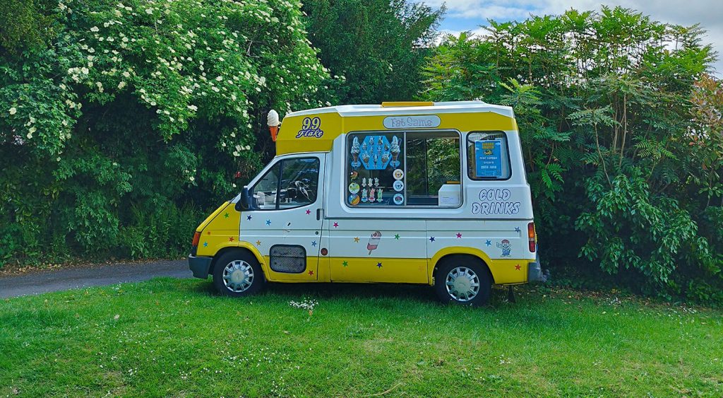 A yellow and white "Fat Sam’s" ice cream van parked against a backdrop of dense green foliage and towering trees under a partly cloudy sky. The serving window is open, with colourful menu stickers displayed, and the words “Cold Drinks” printed on the side. The scene is peaceful, with soft natural light adding to the summery charm.
