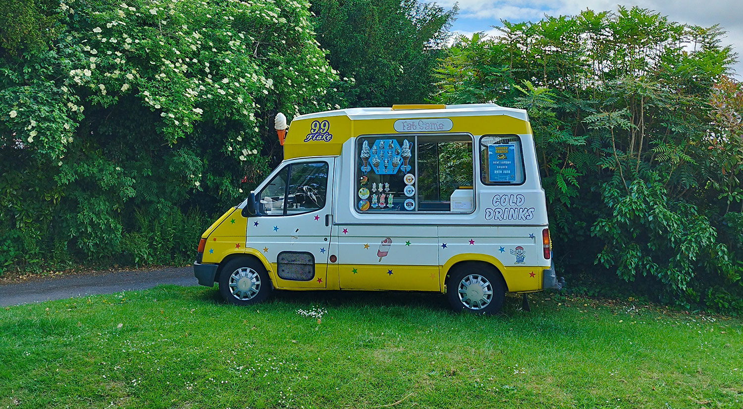 A yellow and white "Fat Sam’s" ice cream van parked against a backdrop of dense green foliage and towering trees under a partly cloudy sky. The serving window is open, with colourful menu stickers displayed, and the words “Cold Drinks” printed on the side. The scene is peaceful, with soft natural light adding to the summery charm.