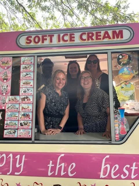 Five smiling women stand inside the pink "Mr Whippy" ice cream van, visible through the large service window. They’re framed by the van's bright pink interior signage advertising various frozen treats. The group appears to be enjoying a fun moment, possibly as part of an event or company visit.