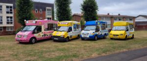 A line-up of four brightly coloured ice cream vans parked on grass in front of residential buildings. The vans are from different brands and eras, including a pink "Mr Whippy", a classic yellow and white "Fat Sam's" with 99 Flake branding, a blue and white van with "Freshly Made Just For You" signage, and a solid yellow van. Each van features distinctive designs and cone sculptures on the roof.