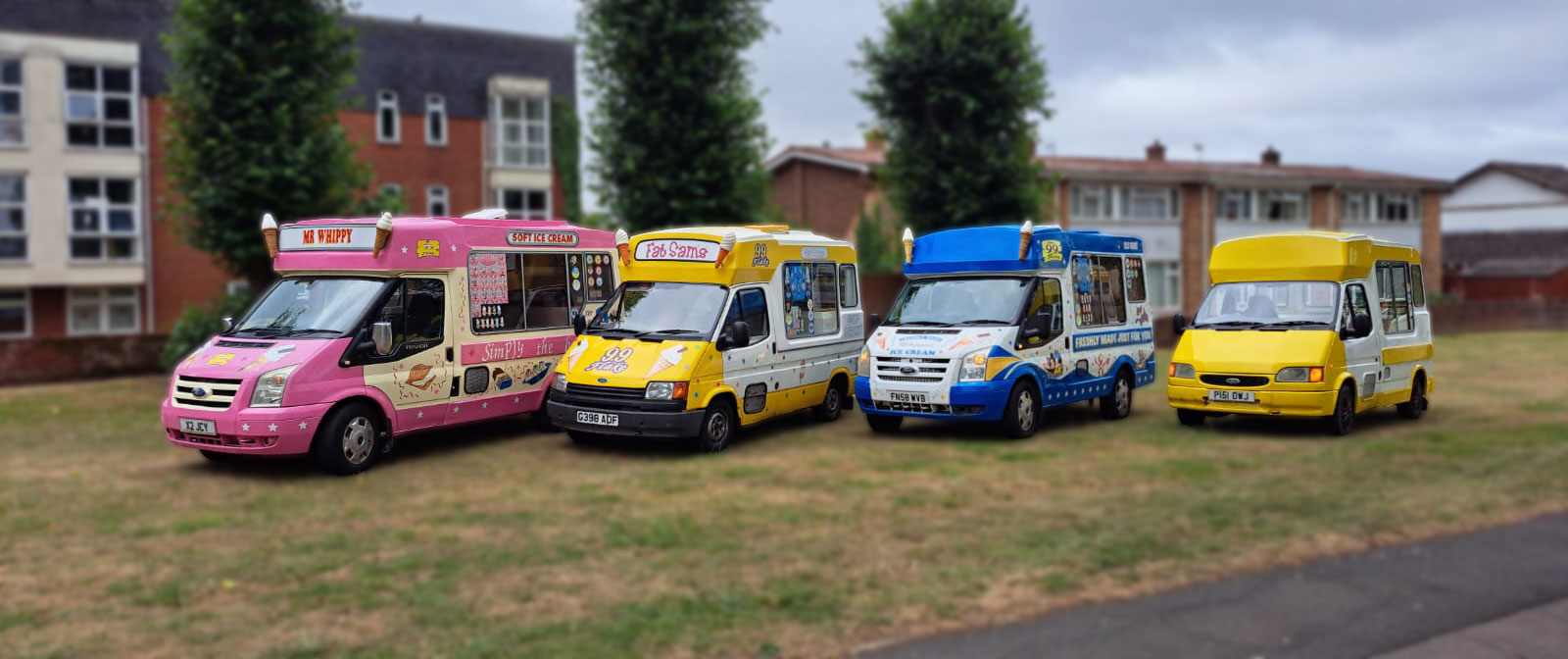A line-up of four brightly coloured ice cream vans parked on grass in front of residential buildings. The vans are from different brands and eras, including a pink "Mr Whippy", a classic yellow and white "Fat Sam's" with 99 Flake branding, a blue and white van with "Freshly Made Just For You" signage, and a solid yellow van. Each van features distinctive designs and cone sculptures on the roof.