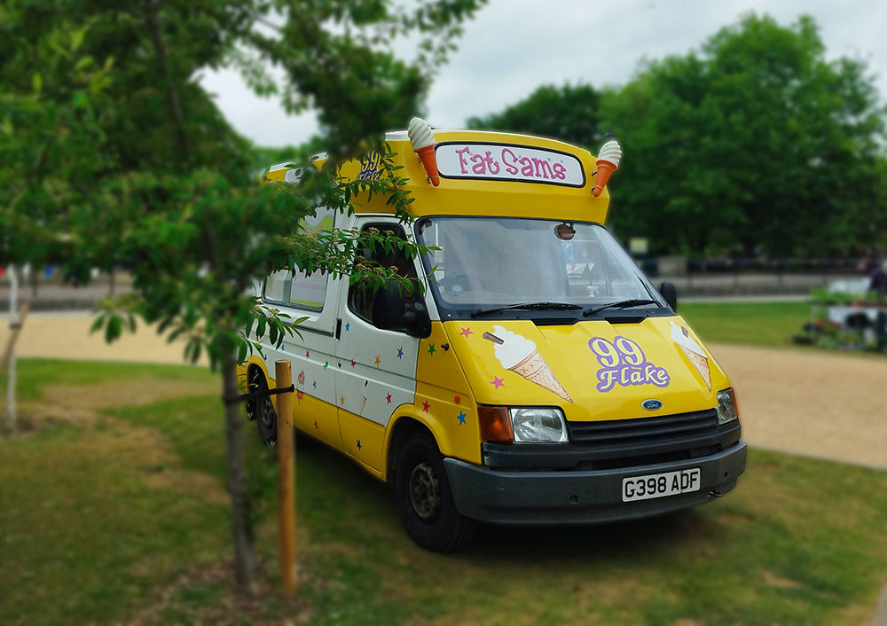 A yellow and white "Fat Sam’s" ice cream van parked against a backdrop of dense green foliage and trees under a partly cloudy sky. The scene is peaceful, with soft natural light adding to the summery charm.