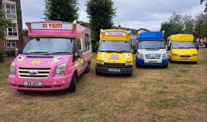 A line-up of four brightly coloured ice cream vans parked on grass in front of residential buildings. The vans are from different brands and eras, including a pink "Mr Whippy", a classic yellow and white "Fat Sam's" with 99 Flake branding, a blue and white van with "Freshly Made Just For You" signage, and a solid yellow van. Each van features distinctive designs and cone sculptures on the roof.