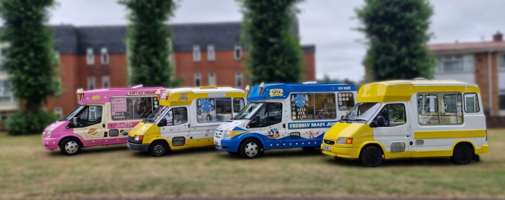 A line-up of four brightly coloured ice cream vans parked on grass in front of residential buildings. The vans are from different brands and eras, including a pink "Mr Whippy", a classic yellow and white "Fat Sam's" with 99 Flake branding, a blue and white van with "Freshly Made Just For You" signage, and a solid yellow van. Each van features distinctive designs and cone sculptures on the roof.