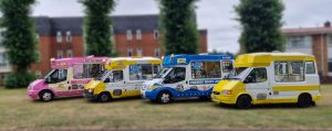 A line-up of four brightly coloured ice cream vans parked on grass in front of residential buildings. The vans are from different brands and eras, including a pink "Mr Whippy", a classic yellow and white "Fat Sam's" with 99 Flake branding, a blue and white van with "Freshly Made Just For You" signage, and a solid yellow van. Each van features distinctive designs and cone sculptures on the roof.