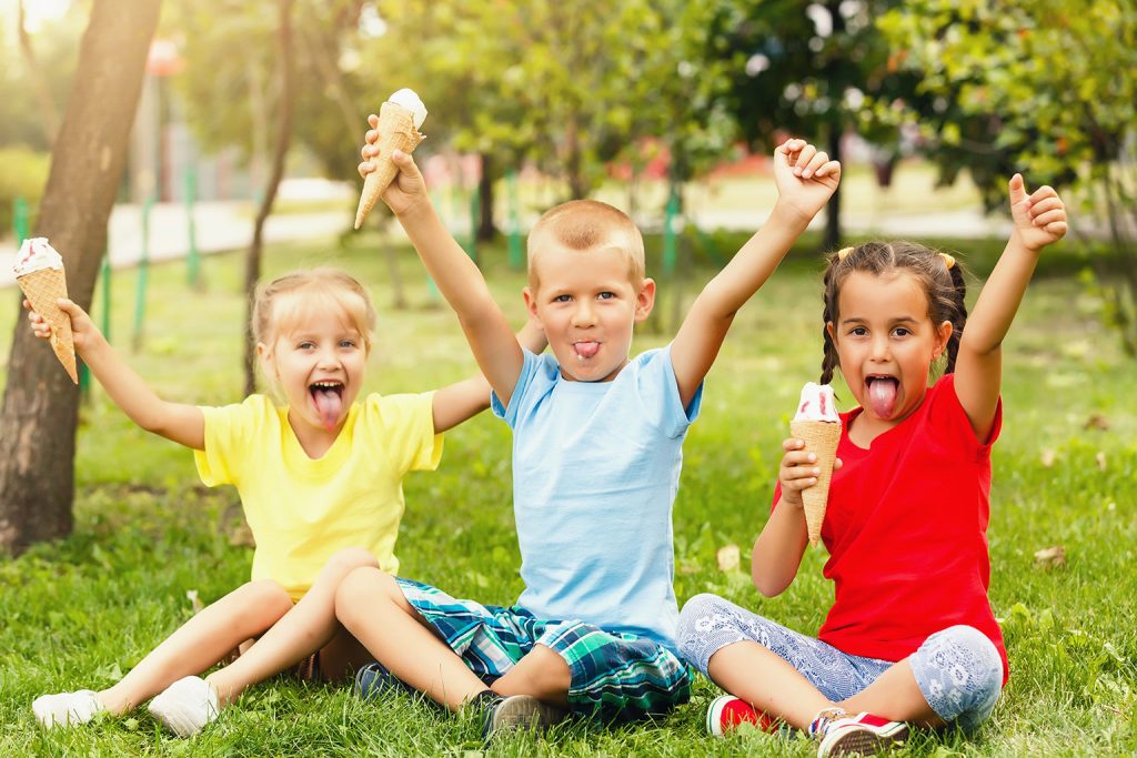 Three young children sitting on grass in a park, smiling with their tongues out and holding ice cream cones, enjoying a sunny summer day.