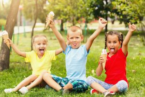 Three young children sitting on grass in a park, smiling with their tongues out and holding ice cream cones, enjoying a sunny summer day.