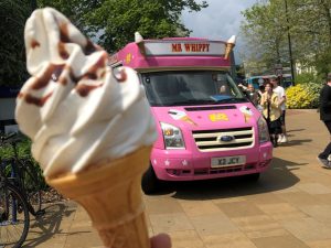 A pink Fat Sam's Ice Cream van at Southampton University serving students ice cream. A whippy ice cream in a cone is being held in front of the ice cream van.
