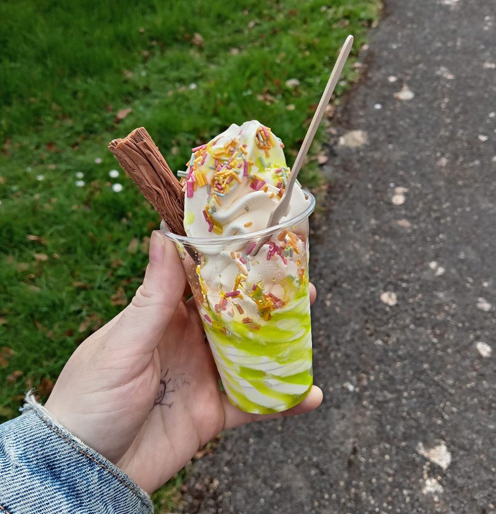 A hand holding a Fat Sam's whipped ice cream cup with lime sauce, colourful sprinkles, and a Cadbury 99 Flake.